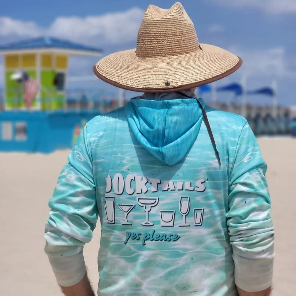 A person standing on a beach wearing a wide-brim straw hat and a turquoise hooded long-sleeve shirt with a white graphic print reading 'DOCKTAILS yes please' on the back. The background shows a blurred beach scene with a lifeguard stand and blue sky.