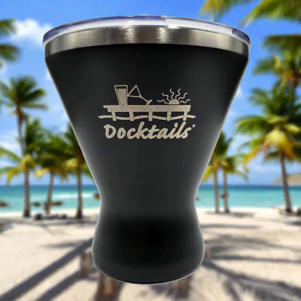 A matte black insulated tumbler with a clear lid, featuring a silver laser-engraved graphic of a cocktail on a dock and the word 'Docktails'. The background is a blurred tropical beach scene with palm trees and blue water.