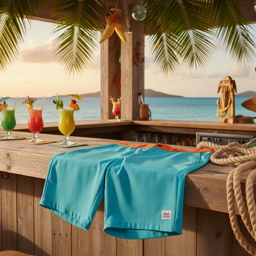A pair of aqua colored men's boardshorts draped over a tiki bar with cocktails next to it and an ocean in the background.