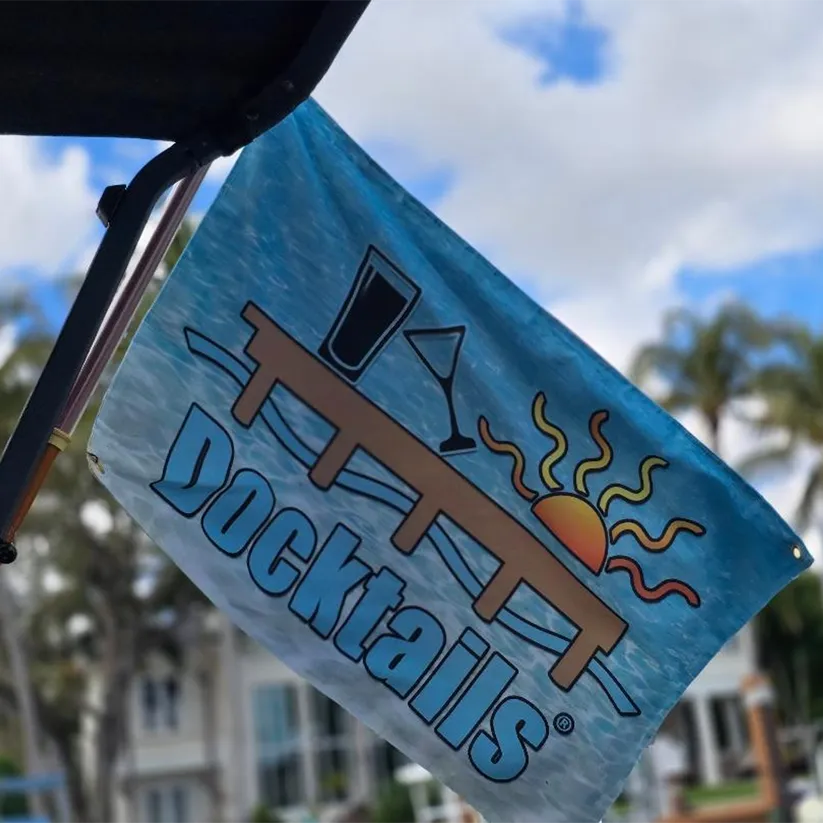Flag hanging at an angle from a boat bimini top. The flag has an aqua colored ocean with a sun, martini glass, pint glass, dock, and the word Docktails.