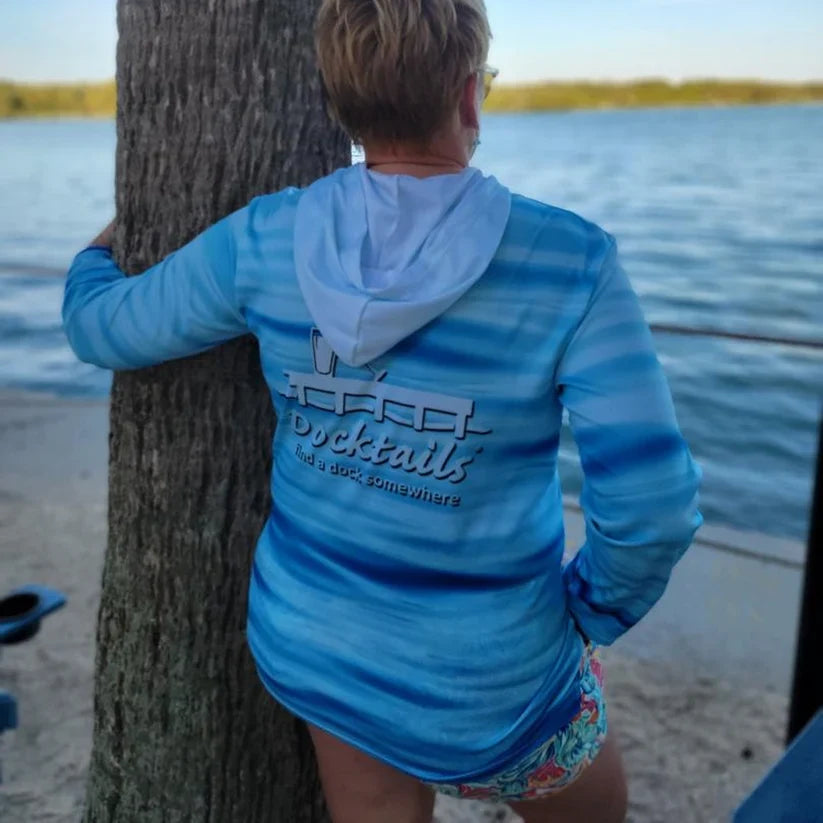 Back of woman standing on a beach at the edge of the water with her arm around a tree. She is wearing a hooded sun shirt with the Docktails logo on it.