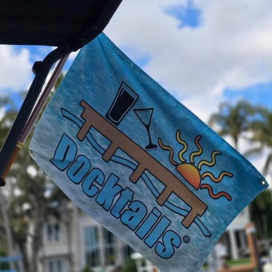 Flag flying at an angle, display the word Docktails, a sun, tilted martini glass, pint glass and a dock, set against an aqua-colored ocean backdrop. The background shows a blurred image of a house.
