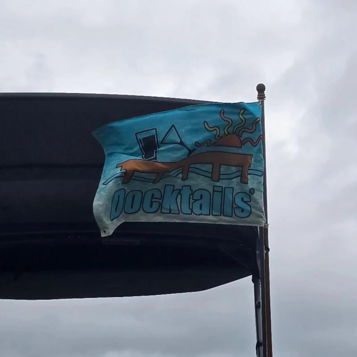 Flag flying from a boat, showing the word Docktails, a sun, tilted martini glass, pint glass and a dock set against an aqua-colored ocean.
