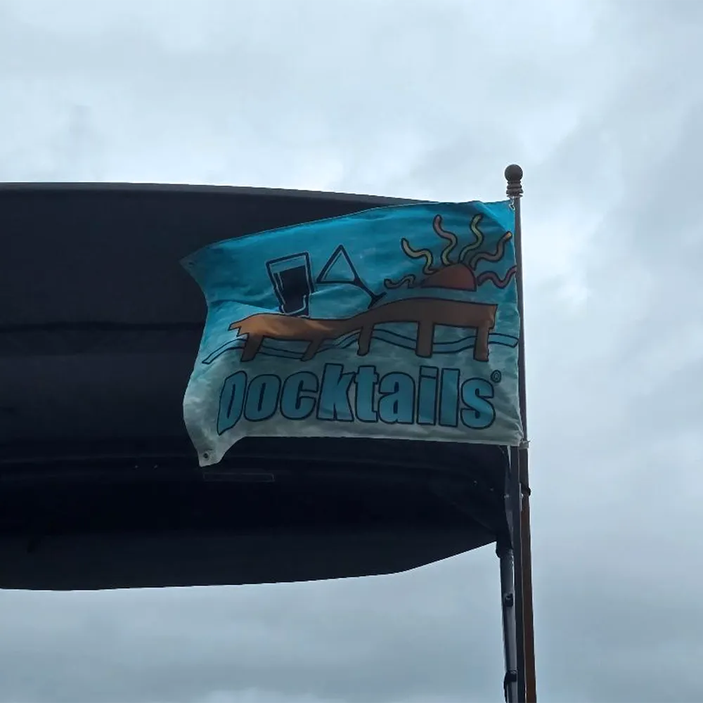 A light blue flag with a 'Docktails' graphic featuring a cocktail glass, beer mug, dock,and sun flying from a boat flag holder under a cloudy sky.