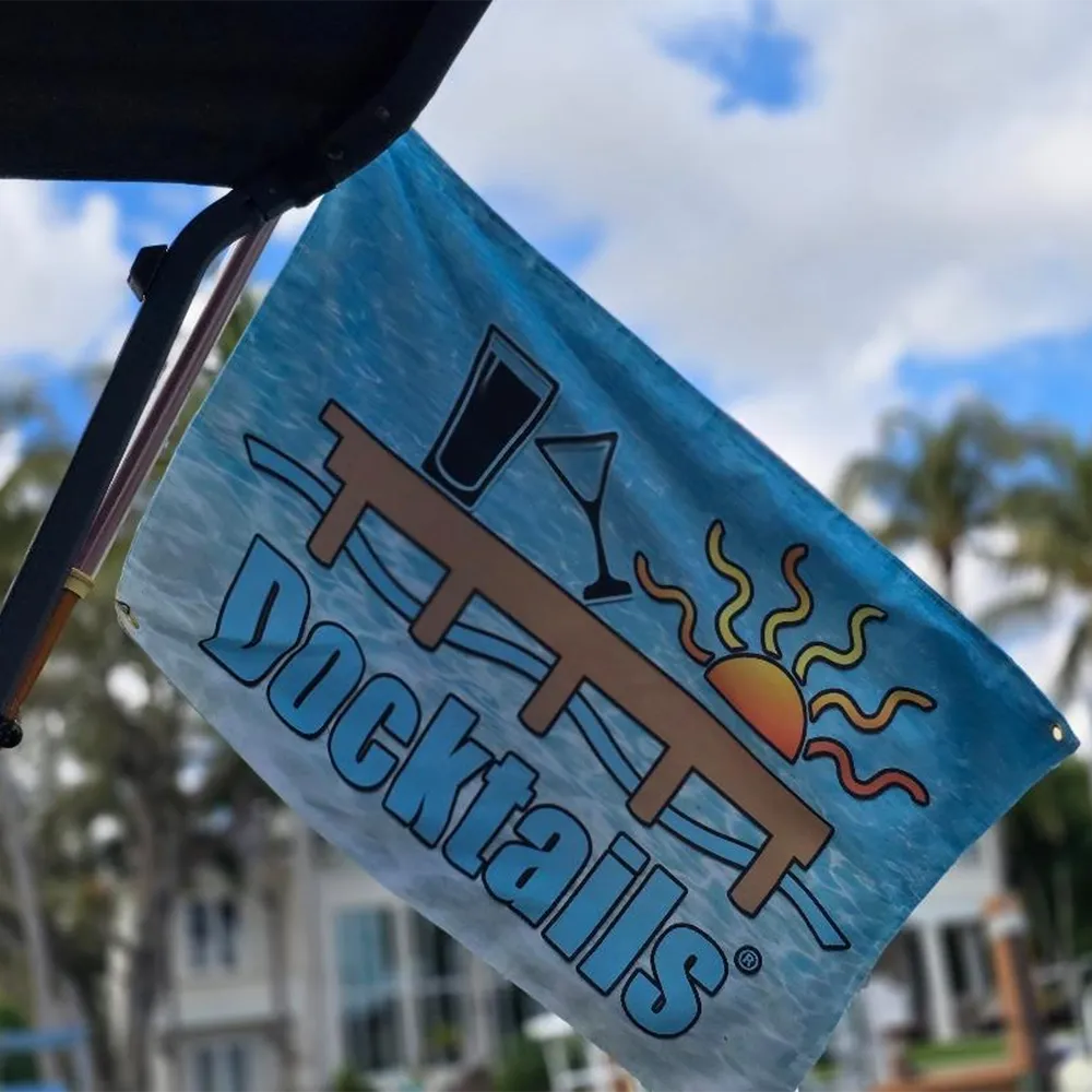 A blue 'Docktails' cocktail-themed flag hanging from a boat or vehicle, with a graphic of a drink, martini glass, dock, and sun, set against a blurred outdoor background of palm trees and sky.