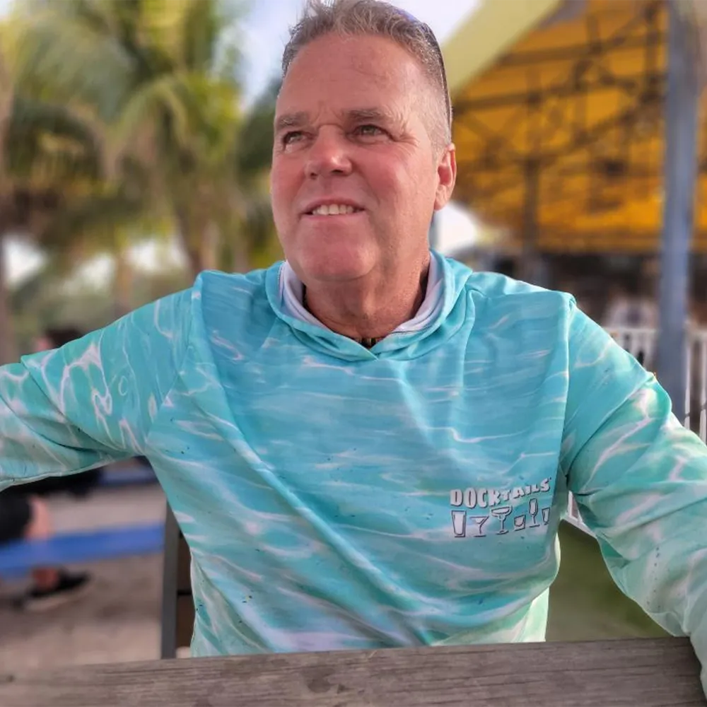 A smiling man wearing a turquoise and white patterned long-sleeve shirt with 'Docktails' printed on the chest, photographed outdoors at a beach bar with blurred palm trees and a yellow structure in the background.