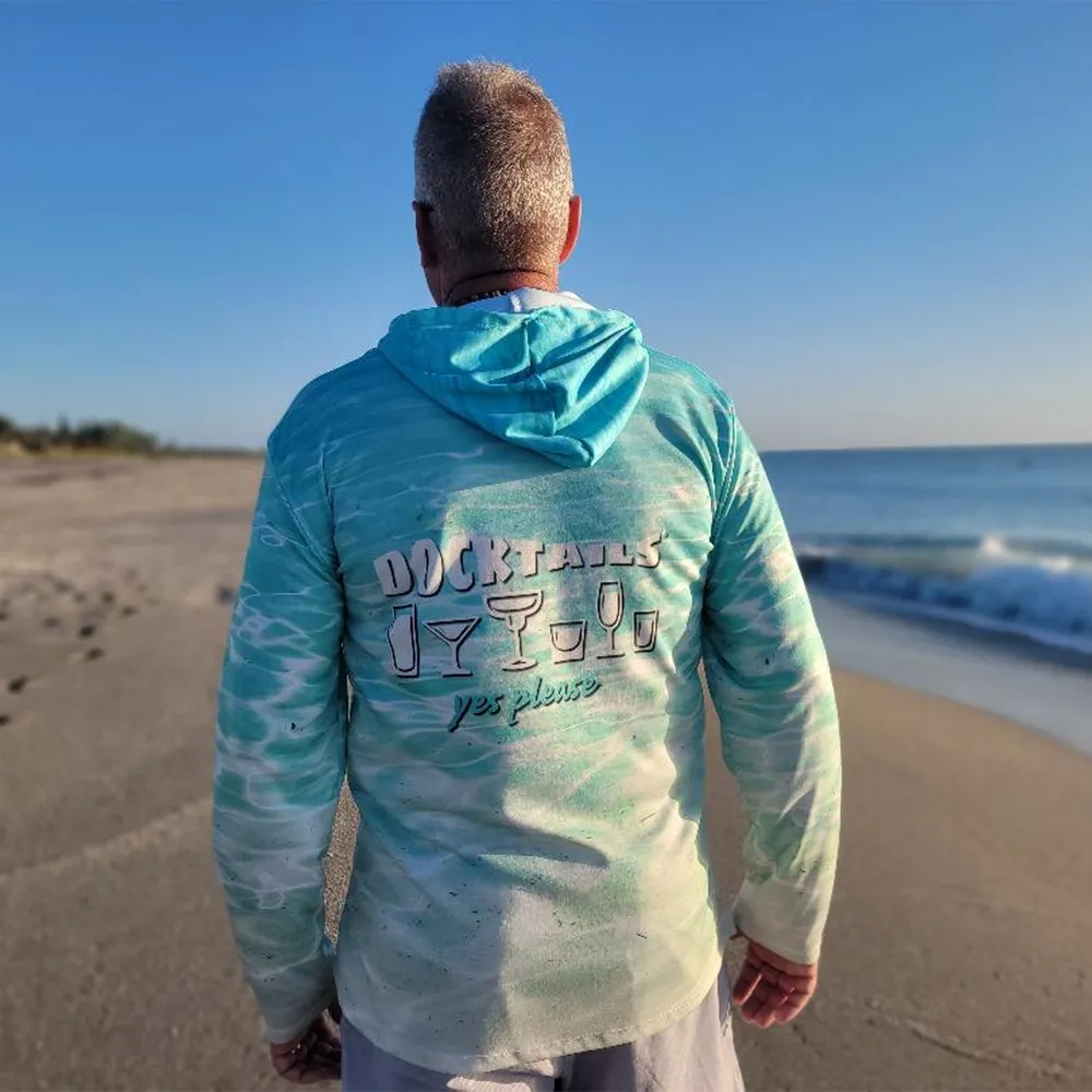 A man standing on a sandy beach with the ocean and blue sky behind him, wearing a turquoise and white hooded long-sleeve shirt with 'Docktails yes please' printed on the back.
