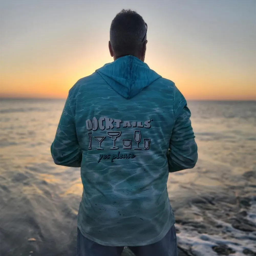 A man stands with his back to the camera at the ocean during sunrise, wearing a turquoise hooded long-sleeve shirt with 'DOCKTAILS yes please' and cocktail glass graphics printed on the back.