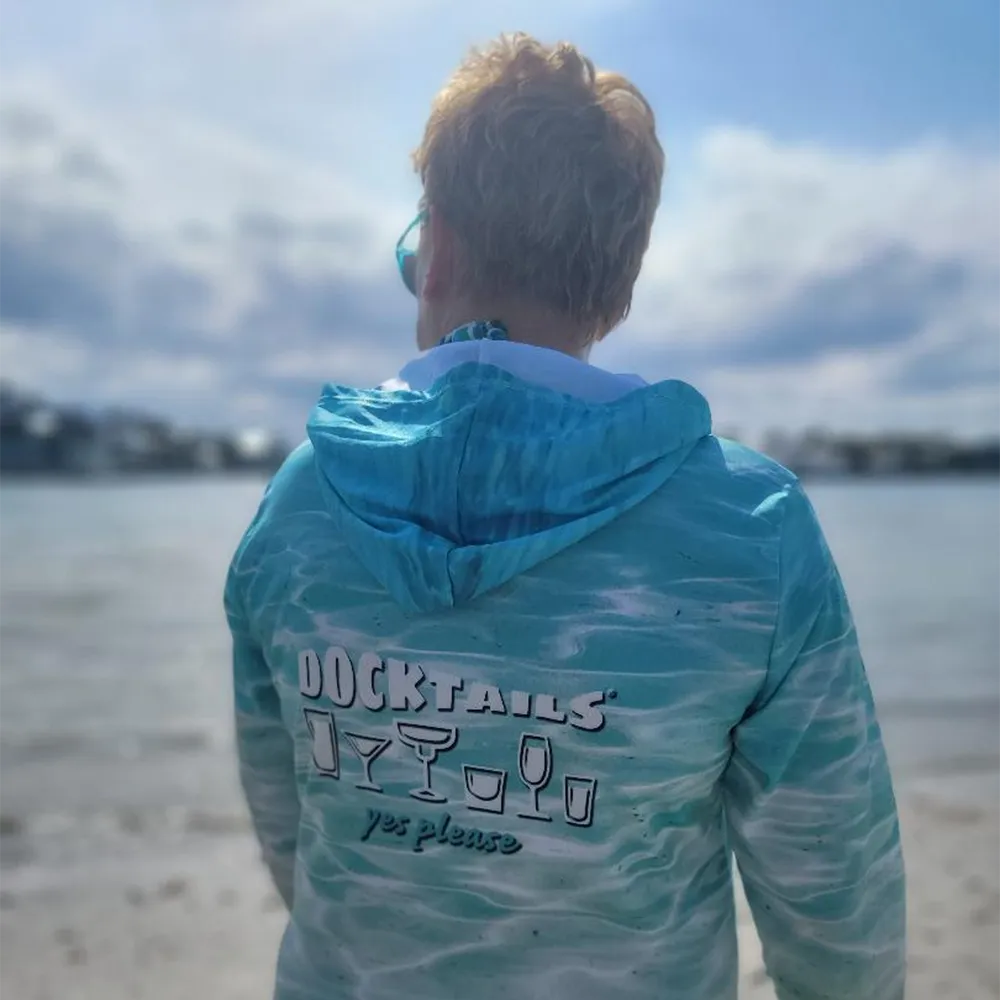 A woman viewed from behind wearing a turquoise and white wavy-patterned hooded shirt with 'DOCKTAILS yes please' printed on the back, standing on a beach with water and a cloudy sky in the background.