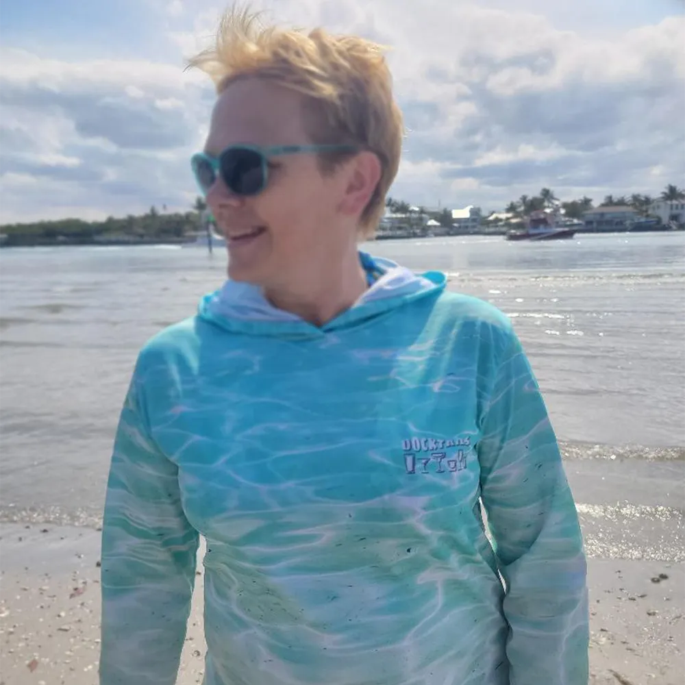 A woman wearing a turquoise and white water-pattern hooded long-sleeve shirt stands on a beach with water and a cloudy sky in the background.