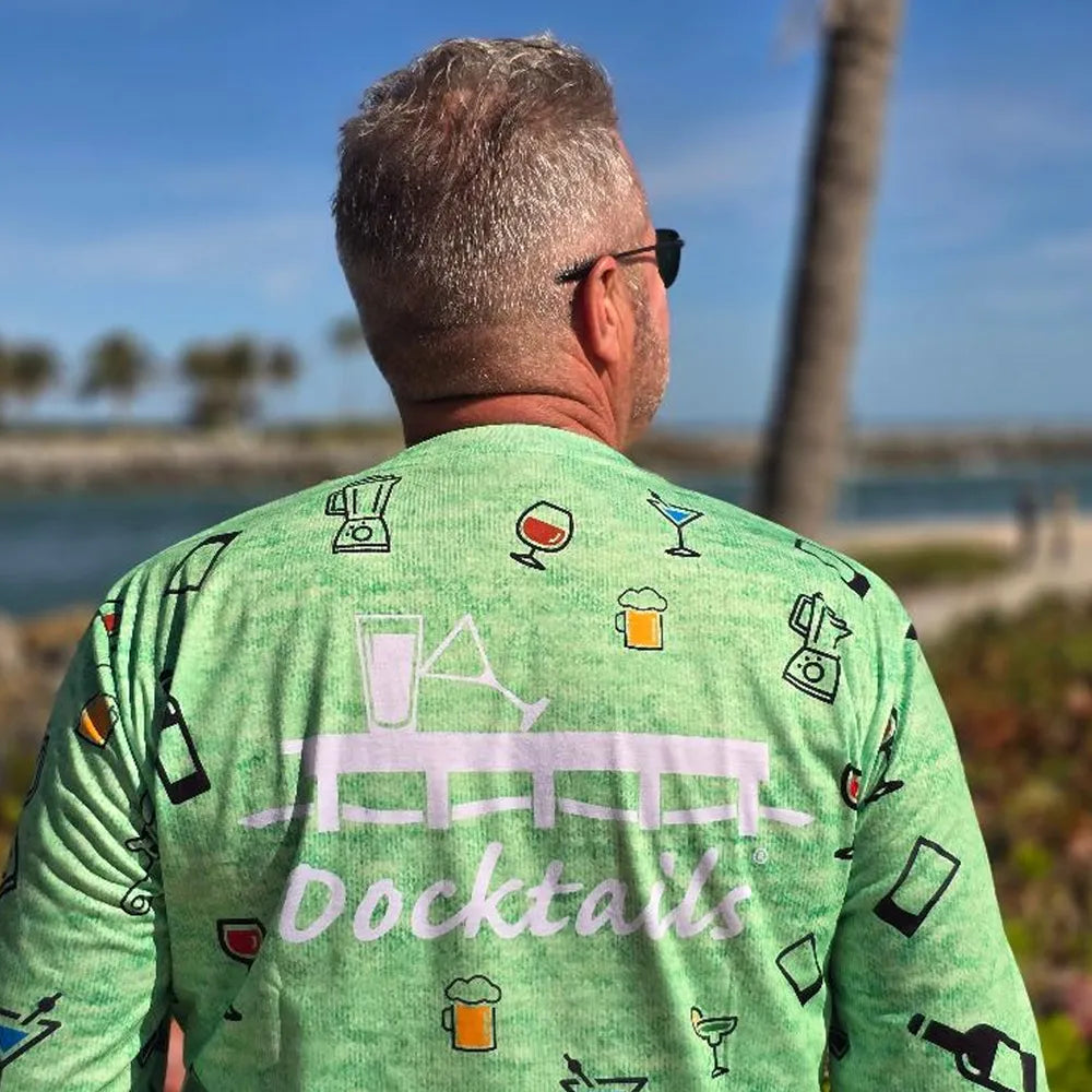 A man viewed from behind wearing a light green long-sleeve shirt with a white 'Docktails' logo and cocktail-themed illustrations across the back, set against a sunny outdoor beach background.