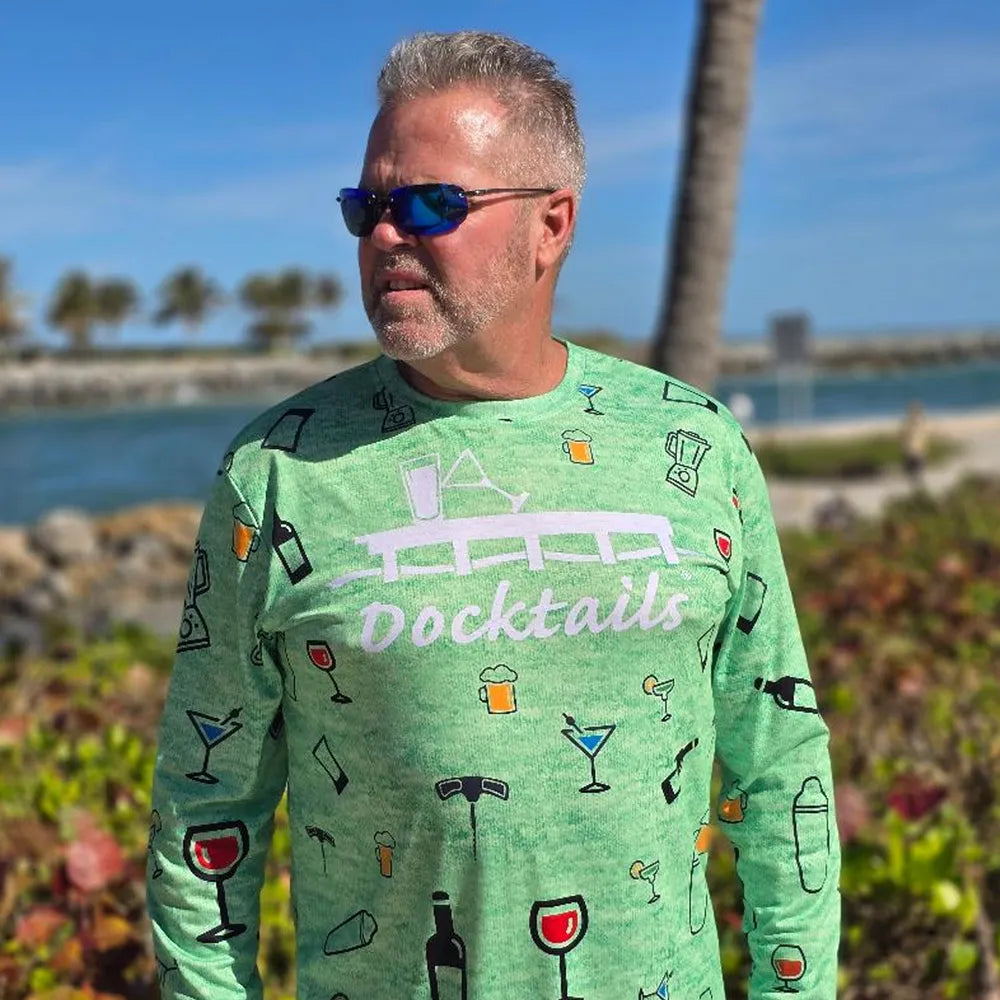 A man wearing a bright green long-sleeve t-shirt with a 'Docktails' cocktail-themed graphic, standing outdoors in front of a beach and palm tree.