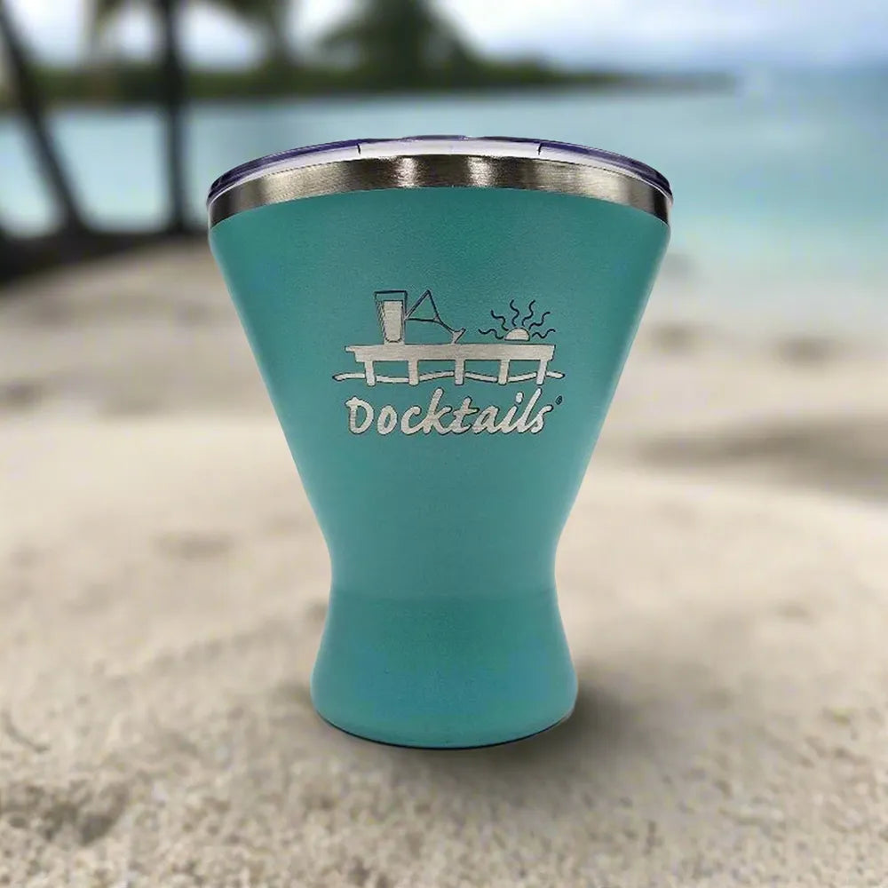 An aqua insulated tumbler with a stainless steel rim and a clear lid, featuring an engraved 'Docktails' graphic on the front, set against a blurred beach background.