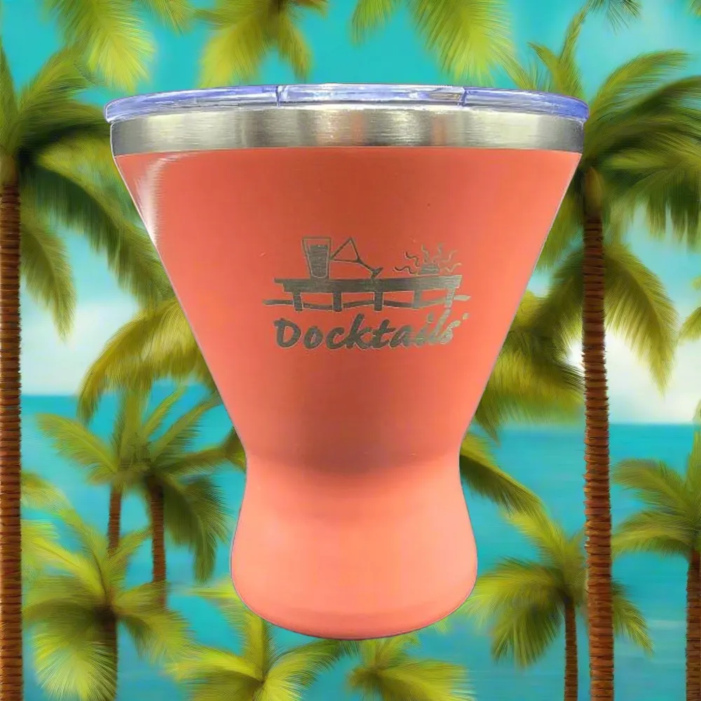 A coral pink insulated tumbler with a clear lid and a silver metal rim, featuring a laser-engraved 'Docktails' cocktail graphic. The tumbler is set against a tropical beach background with palm trees and a blue ocean.
