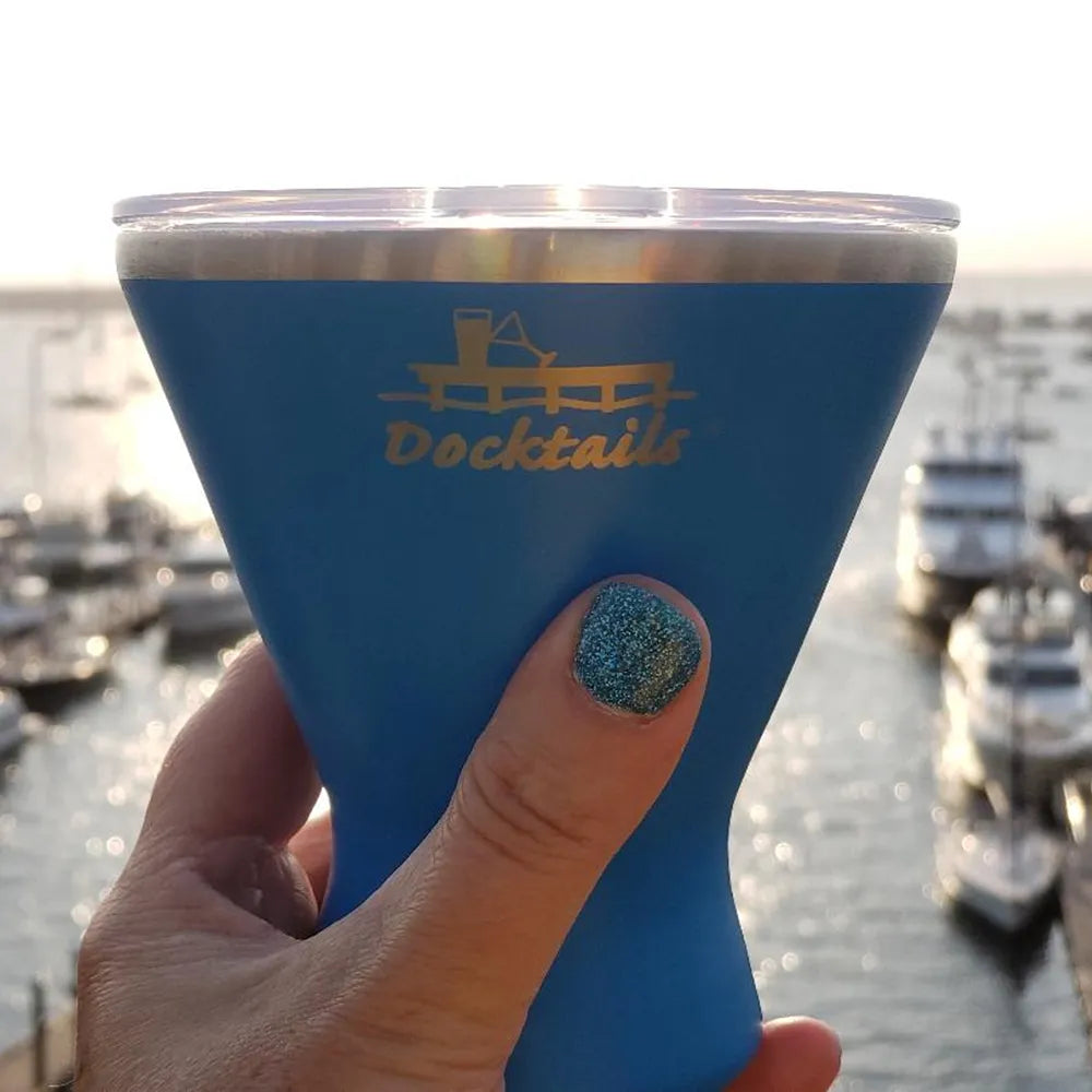 A hand with glittery teal nail polish holds a blue insulated tumbler with a clear lid. The tumbler is engraved with a dock logo and the word 'Docktails'.