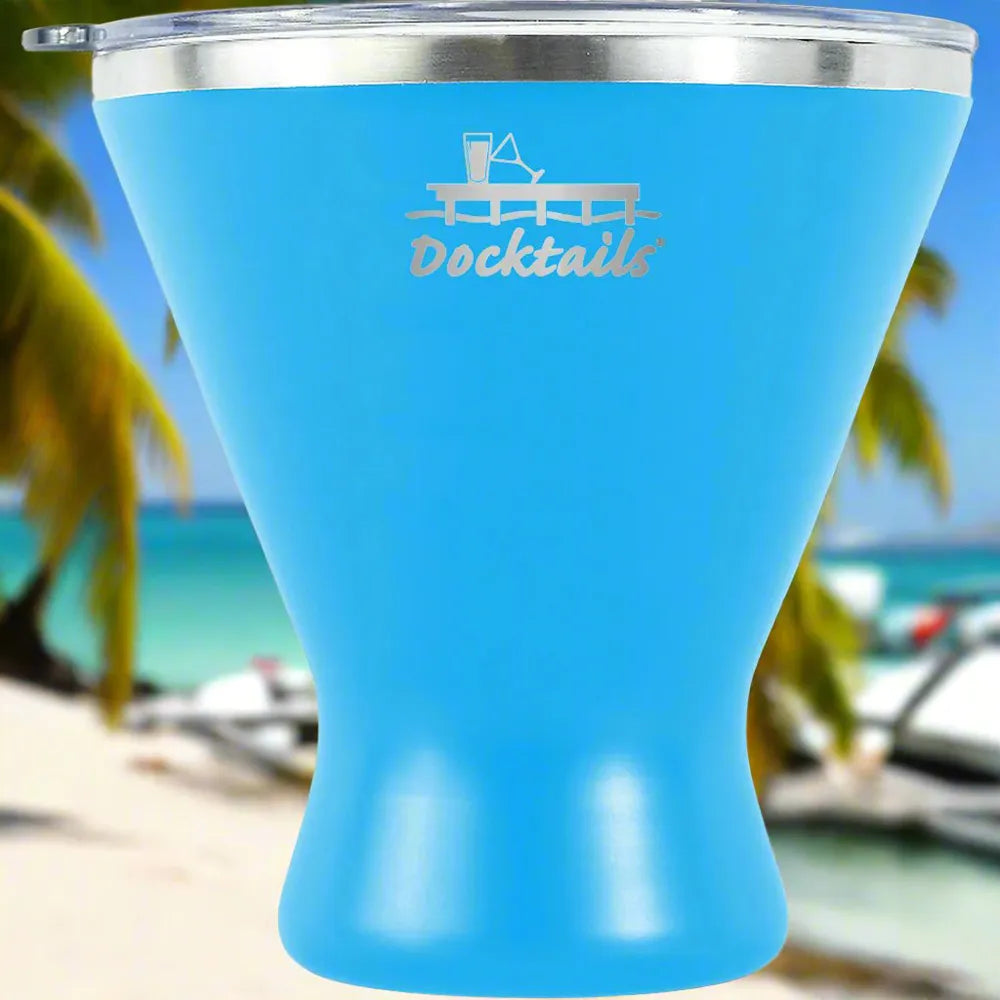 A bright blue insulated tumbler with a silver rim and clear lid, featuring an engraved 'Docktails' graphic with a cocktail glass and pint glass on a dock. The background is a blurred tropical beach scene with palm trees and ocean.