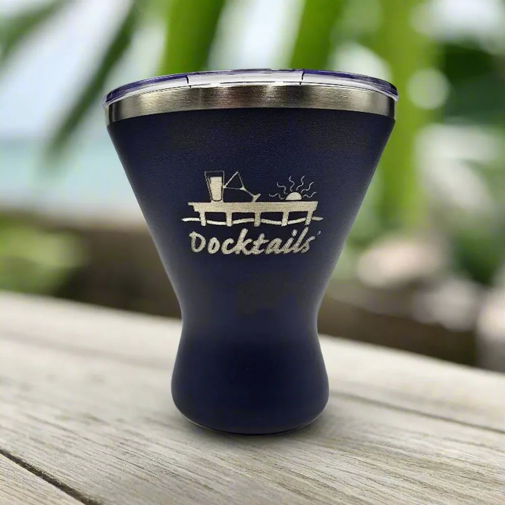 A navy blue insulated tumbler with a stainless steel rim and a lid, featuring an engraved graphic of a cocktail and the word 'Docktails' on the front, set on a wooden surface with a blurred outdoor background.
