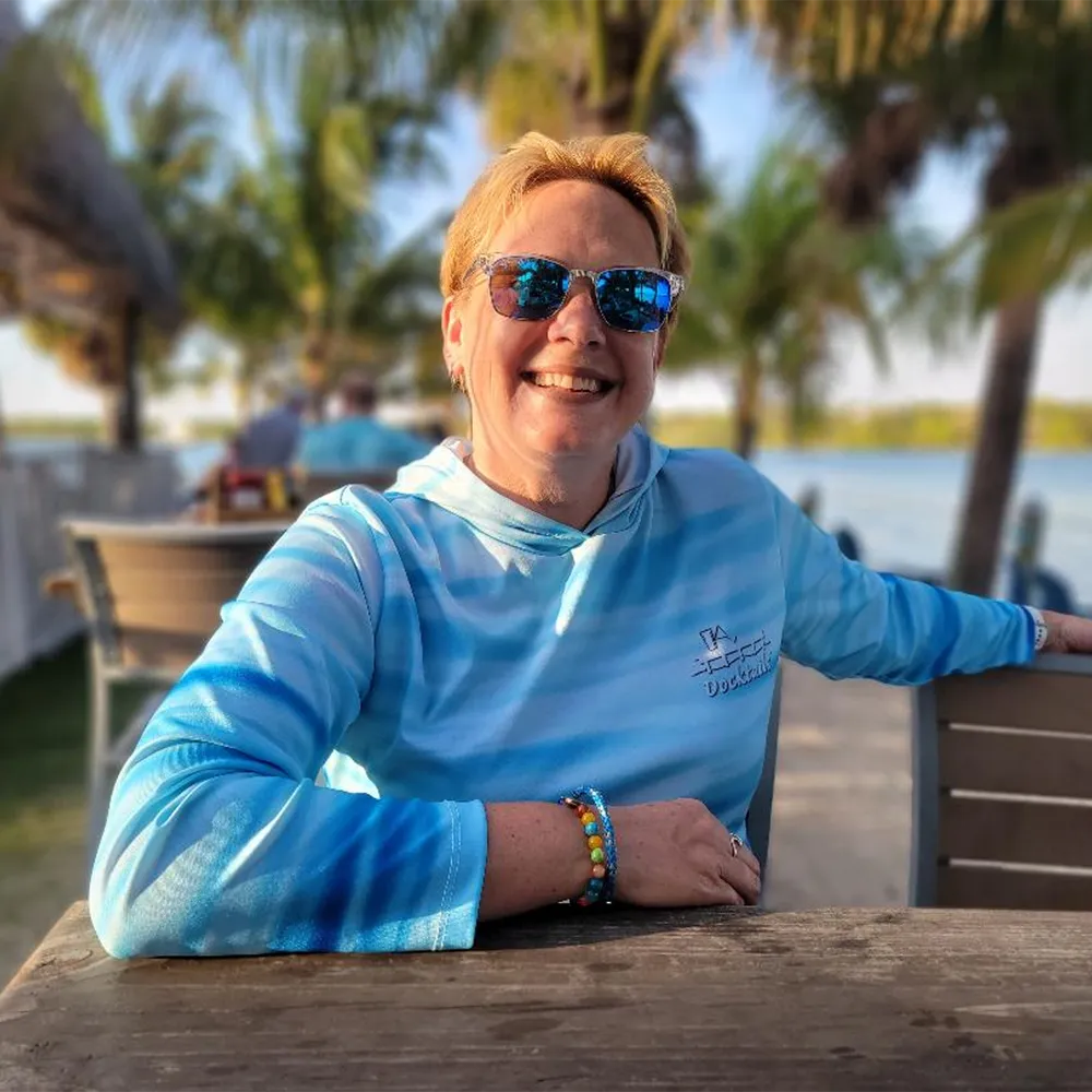 A smiling woman with short blonde hair and blue mirrored sunglasses sits at an outdoor table by the water, wearing a blue and white long-sleeve hooded shirt with a small white logo on the left chest.