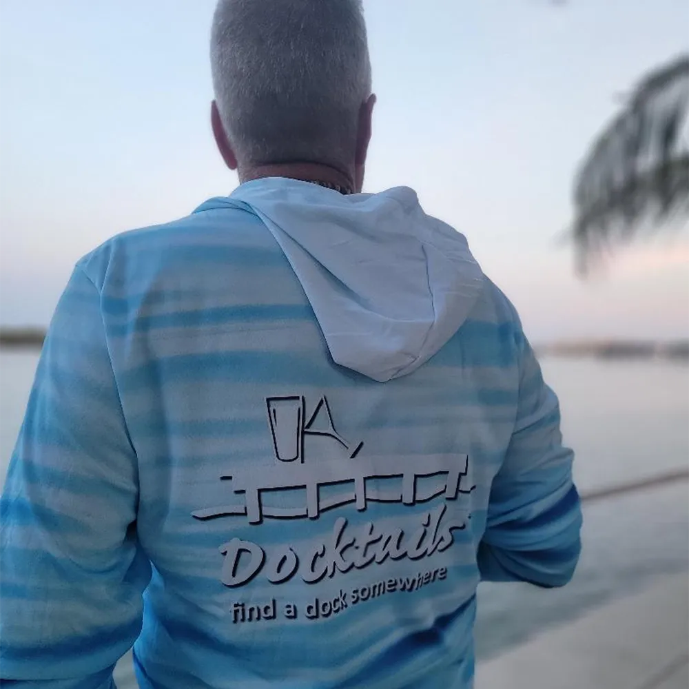 A man viewed from behind wearing a light blue and white striped hoodie with 'Docktails' printed on the back, set against a soft-focus outdoor background near water.