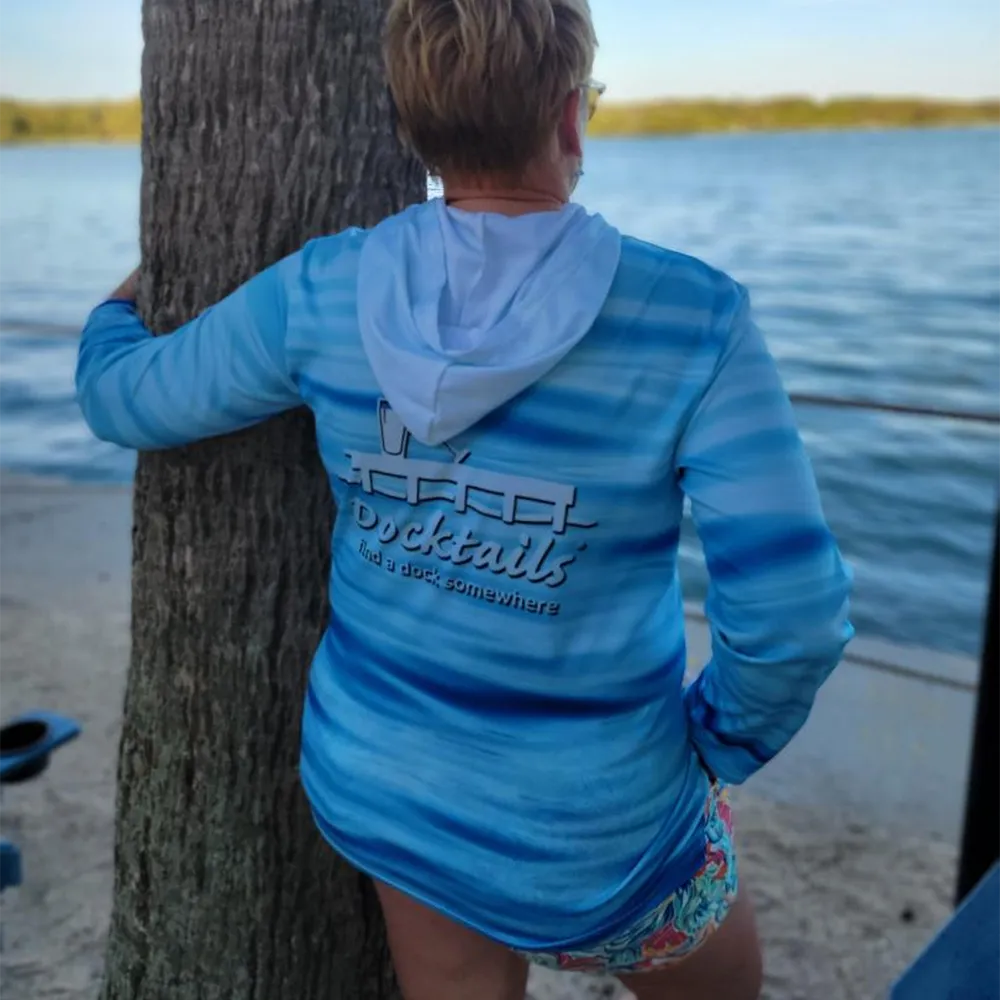 A woman stands outdoors by the water, seen from behind, wearing a blue and white wave patterned hooded long-sleeve shirt with a white graphic logo that reads 'Docktails' and 'find a dock somewhere'.