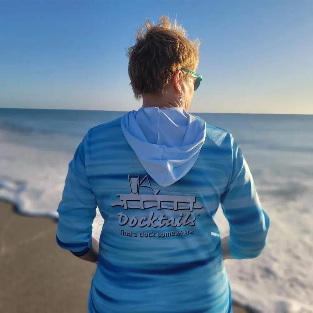 A woman viewed from behind wearing a blue and light blue wave-patterned long-sleeve hoodie with a hood, standing on a beach with the ocean and sky in the background. The back of the hoodie features a graphic with the text 'Docktails' and 'find a dock somewhere'.