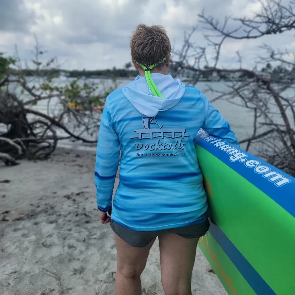 A woman stands on a sandy beach facing the water, wearing a light blue and white hooded long-sleeve shirt with a large 'Docktails' logo on the back, and holding a green and blue paddleboard.