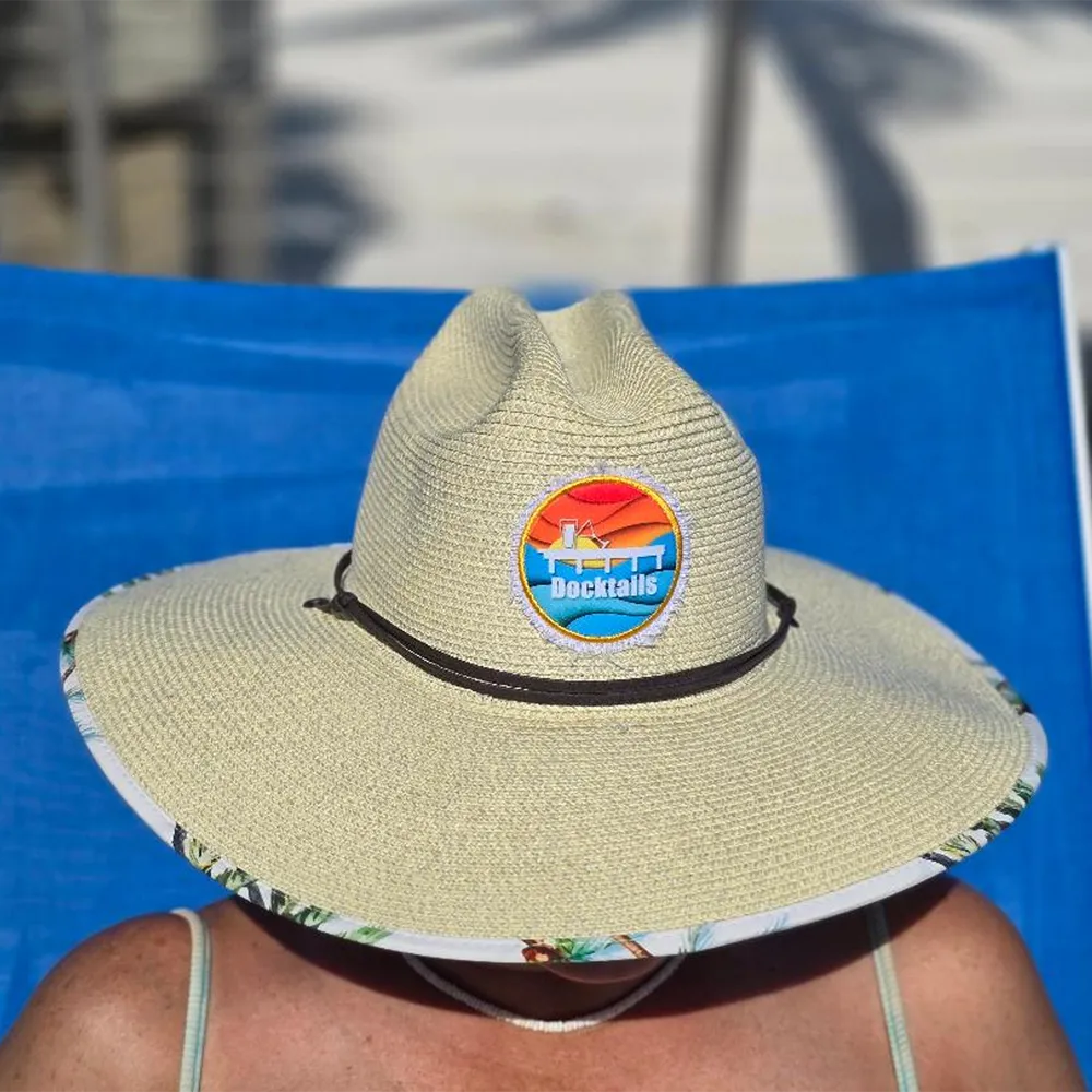 Woman sitting on a blue chair wearing a beige wide brim sun hat with a Docktails logo patch on it. The hat hides the woman's face.
