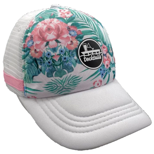 A white trucker hat with a tropical floral and palm leaf pattern on the front panel, a white mesh side panel with a pink fabric band, and a black circular 'Docktails' logo patch on the crown.