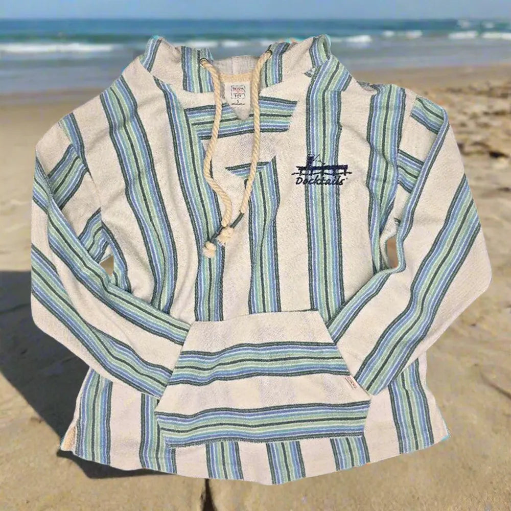 A long-sleeve striped hoodie with a drawstring neckline and front pouch pocket, laid flat on a sandy beach with the ocean in the background. The garment features vertical stripes in blue and green on an off-white base, with a small Docktails logo on the left chest.