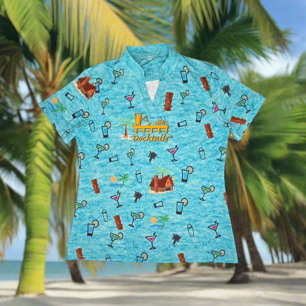 A light blue short-sleeve women's collared polo shirt with a tropical cocktail-themed print, displayed against a blurred beach and palm tree background.