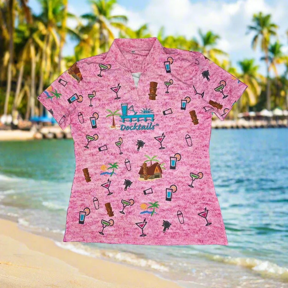 A women's pink short-sleeve collared polo shirt with an all-over tropical cocktail and beach-themed print, shown against a blurred beach and palm tree background.